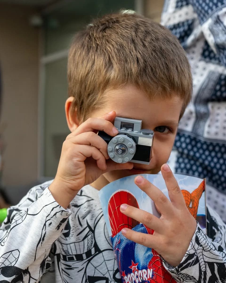A young boy holds a toy brick camera up to his face so it covers one eye; his other eye looks toward the lens. He wears a white sweatshirt with a black comic print and holds a printed popcorn box, and a person in a navy and white patterned top stands at the right edge of the frame.