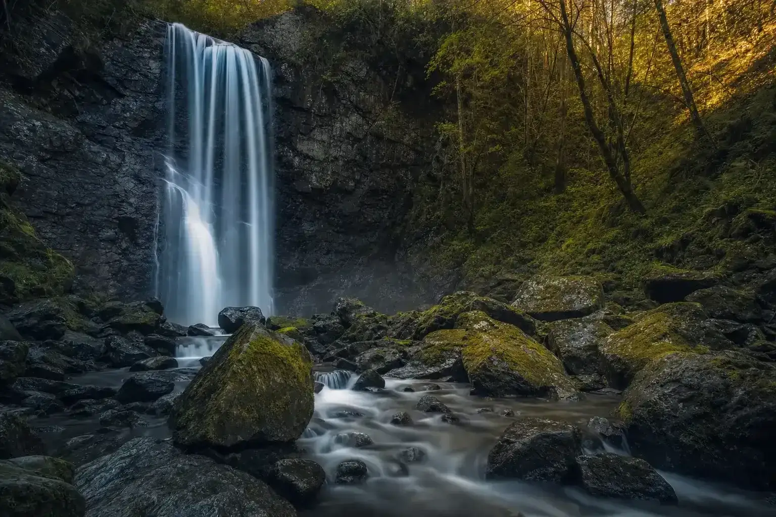 AI-generated image of a forest waterfall on a long exposure: the falling water rendered as silky luminous veils over dark rock while every mossy boulder in the stream below stays tack-sharp, warm evening light catching the trees above.