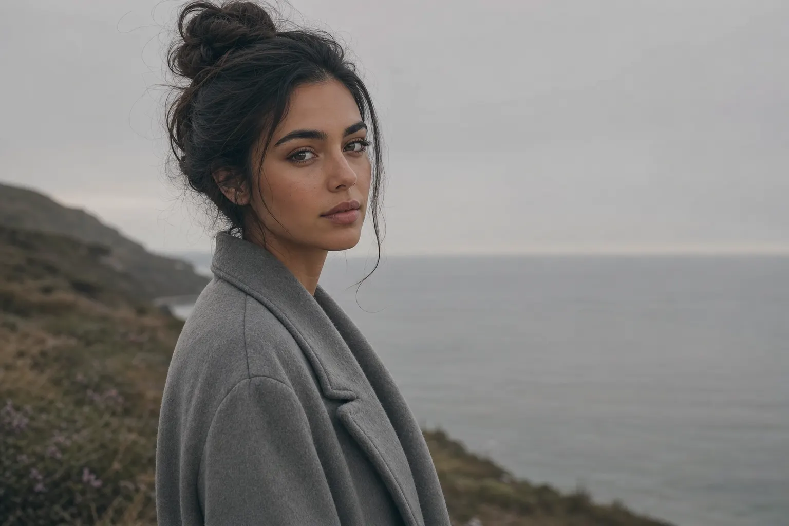 AI-generated image of a dark-haired woman in a soft grey wool coat on a heather clifftop on an overcast day, her face in gentle three-quarter view in the even light, a calm pale-grey sea and soft flat sky behind her.
